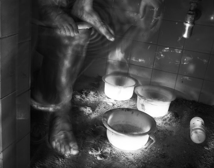 This black and white photograph shows an old bathroom. The floor is covered in a thick layer of dust and dirt. The feet of a ghostly figures sitting behind a wall are floating off to the left.