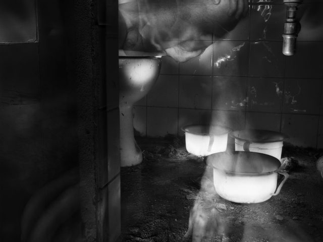 This black and white photograph shows the dusty interior of an old bathroom. The floor is covered in a thick layer of dirt. Three white bowls sit in it. The legs of a naked man rise up on the left.