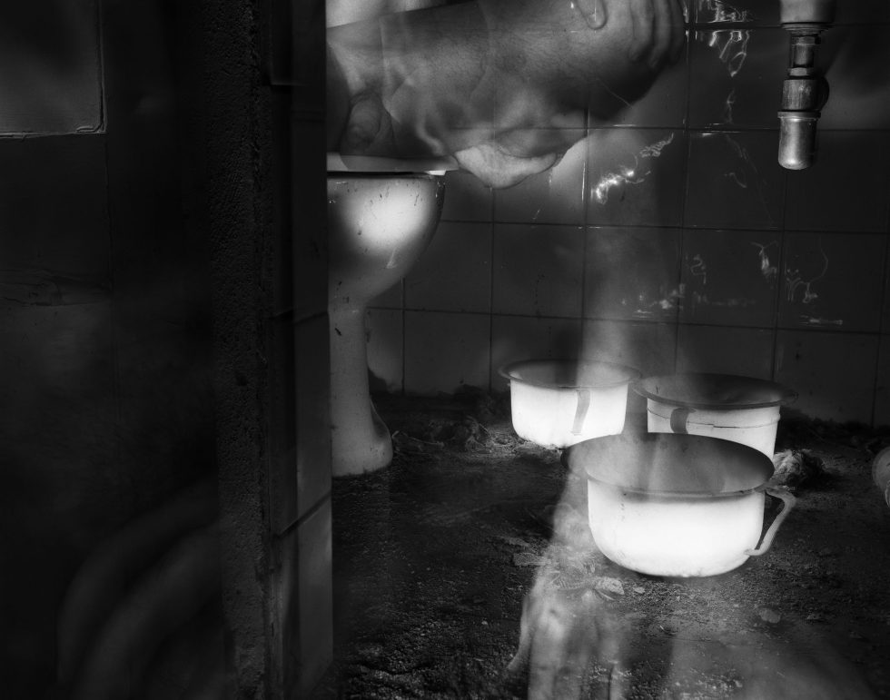 This black and white photograph shows the dusty interior of an old bathroom. The floor is covered in a thick layer of dirt. Three white bowls sit in it. The legs of a naked man rise up on the left.