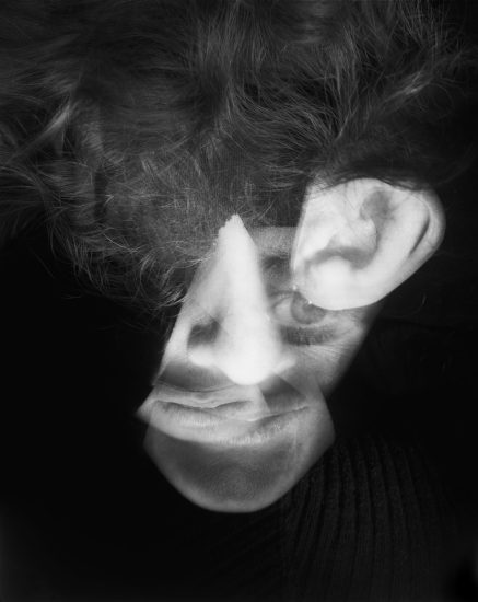 This image shows a black and white close up go a man's face. A cut out of a human ear floats beside his face, partly hidden in shadows.