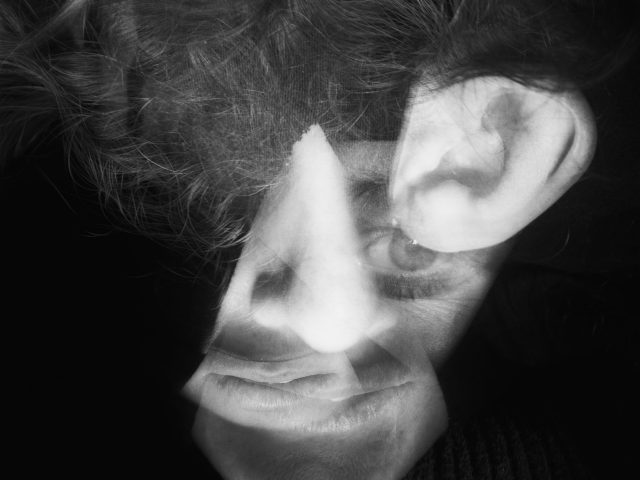 This image shows a black and white close up go a man's face. A cut out of a human ear floats beside his face, partly hidden in shadows.