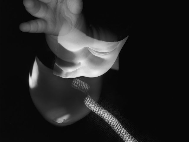 This black and white image shows a ghostly hand reaching outwards. A thick chord snakes upwards.