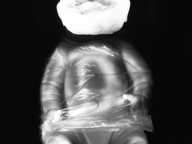 This black and white photograph shows a bright white doll, its face overexposed and featureless. Its body is wrapped in plastic.
