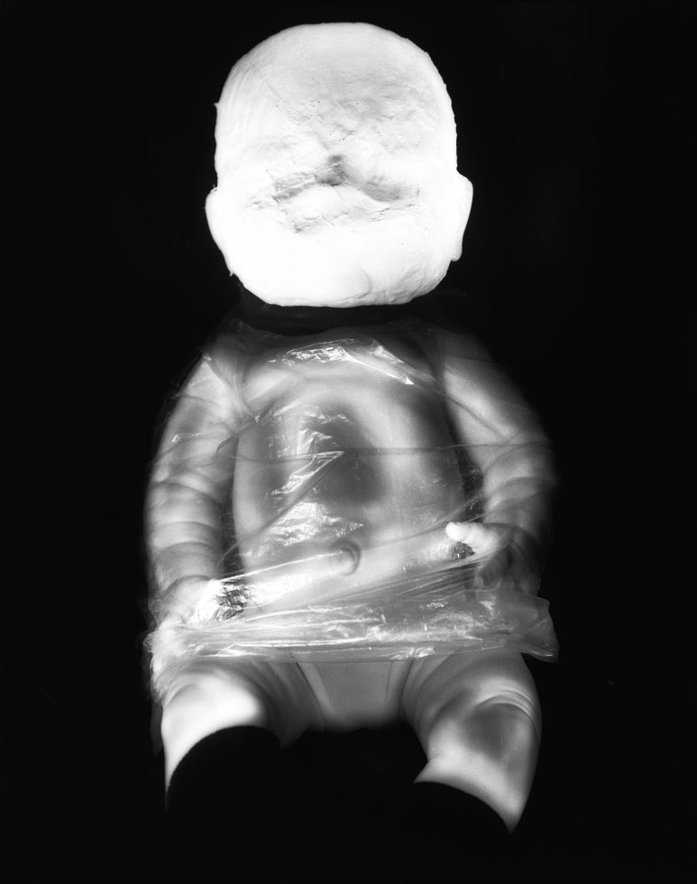 This black and white photograph shows a bright white doll, its face overexposed and featureless. Its body is wrapped in plastic.