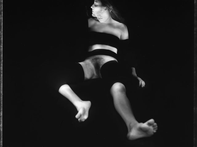 This black and white photograph shows a woman sitting on the ground. She leans into the darkness behind her. She is naked from the waist down.