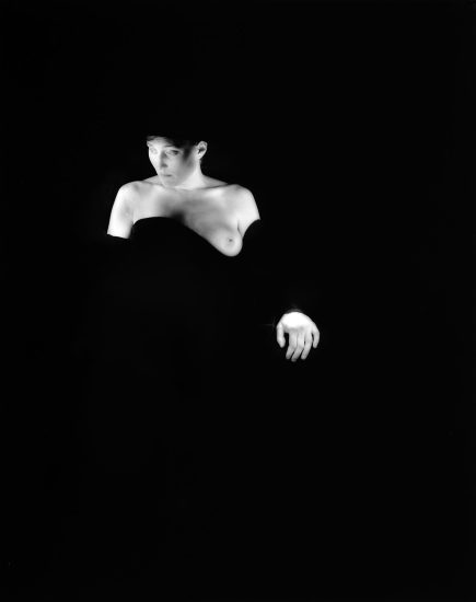 This black and white photograph shows a woman against a black background. Her skin is bright white and she is partially obscured by a black cloth.