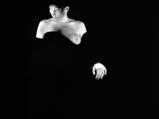 This black and white photograph shows a woman against a black background. Her skin is bright white and she is partially obscured by a black cloth.