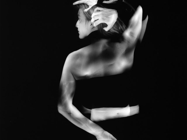 This black and white photograph shows the back of a woman against a black background. Her arms reach above her head and move down her back. They are blurred with motion.