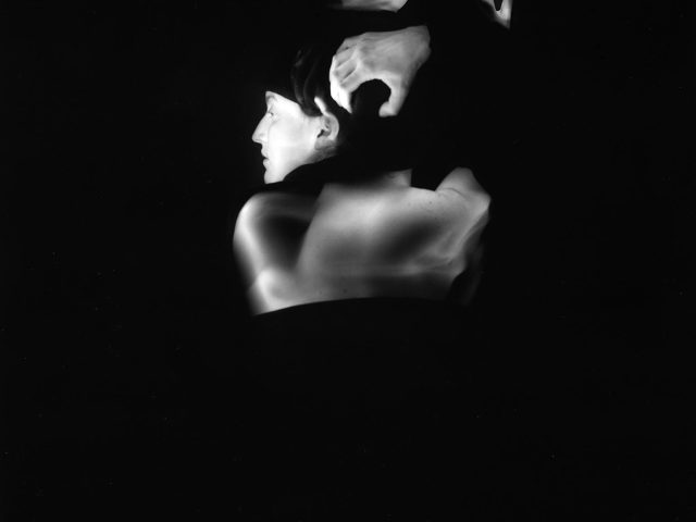 This black and white photograph shows a woman, seen from the back, who appears to have more than two arms. The arms reach above her head and run across her back. They are blurred with motion.