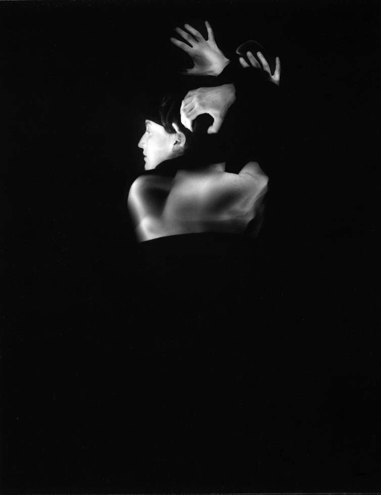 This black and white photograph shows a woman, seen from the back, who appears to have more than two arms. The arms reach above her head and run across her back. They are blurred with motion.