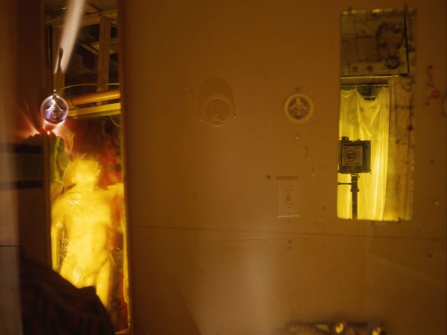 This photograph shows the yellow, glowing walls of a bedroom space where a human figure is seen through a doorway off to the left.