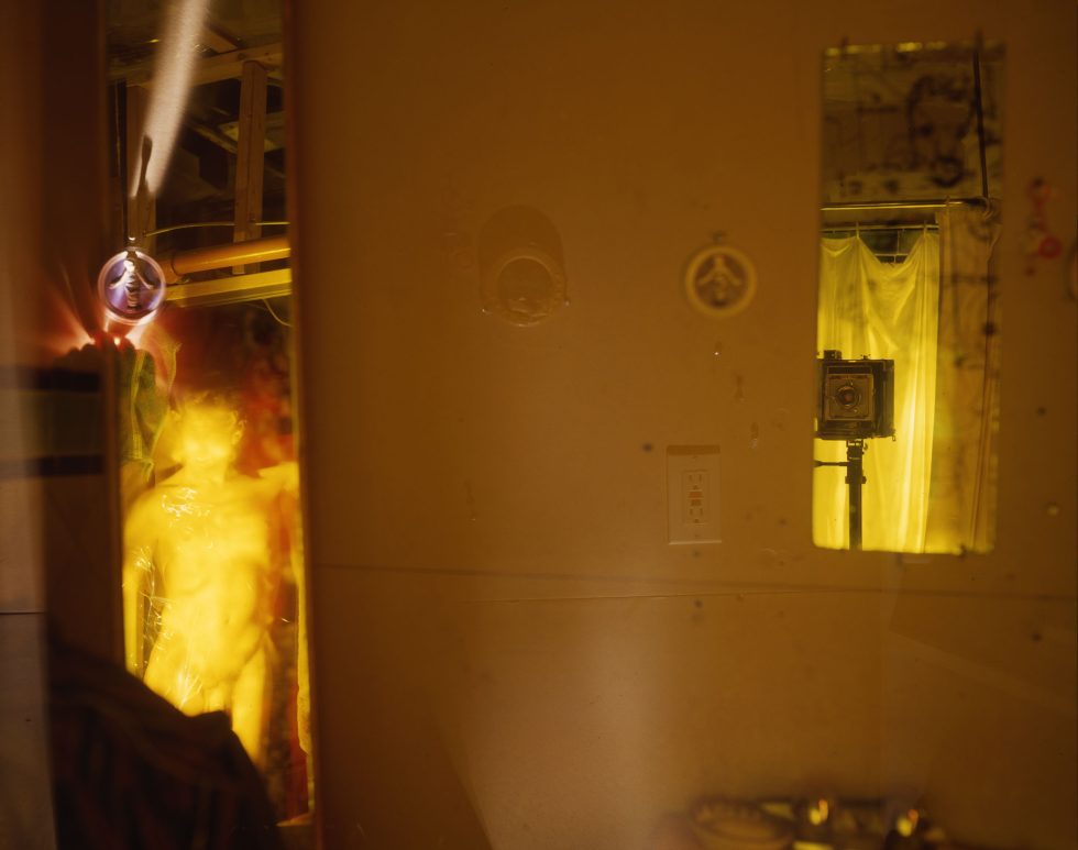 This photograph shows the yellow, glowing walls of a bedroom space where a human figure is seen through a doorway off to the left.
