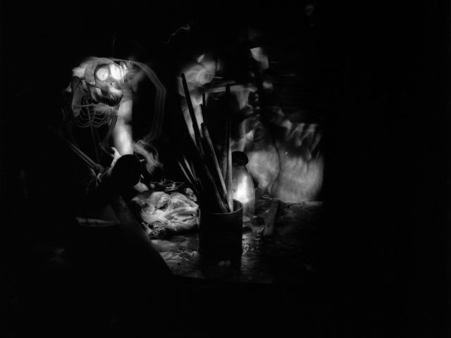 This black and white photograph shows the skeleton of a monkey on a table with a pot of paint brushes.