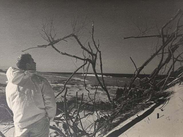 In this photograph, a man on a beach is wearing a puffy coat.