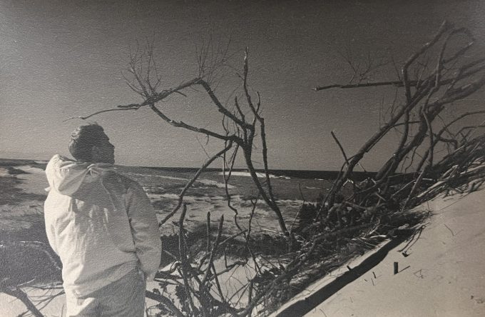 In this photograph, a man on a beach is wearing a puffy coat.