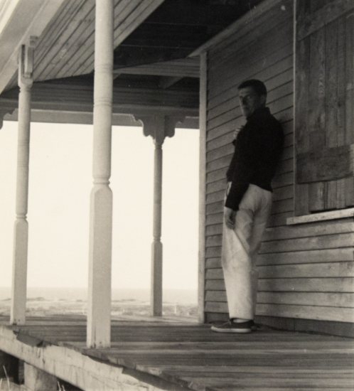 This black and white photograph shows a man standing on an old porch on the beach.
