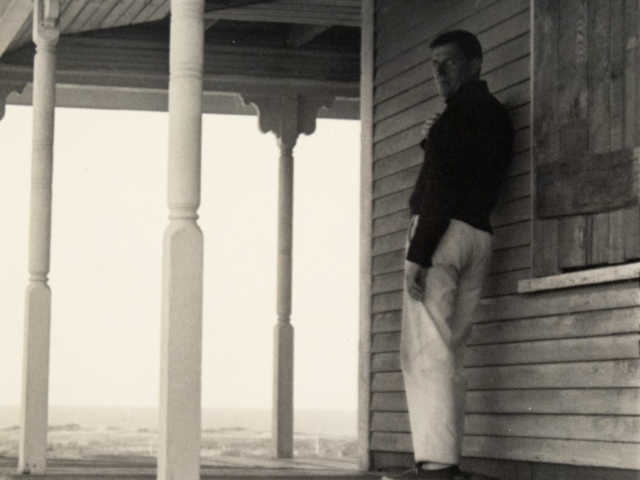 This black and white photograph shows a man standing on an old porch on the beach.