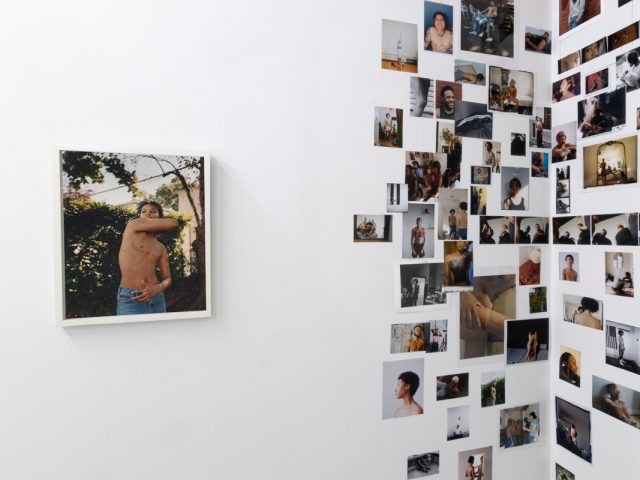 This image shows a collage of images on the walls of a gallery, placed next to a single framed image of a shirtless person with their arm wrapped around their body.