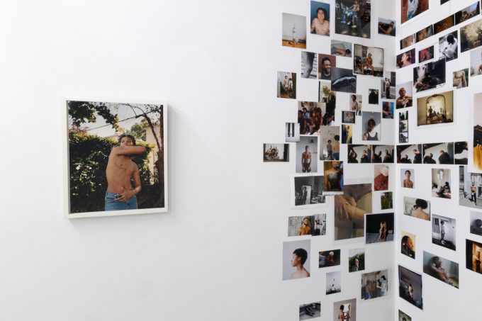 This image shows a collage of images on the walls of a gallery, placed next to a single framed image of a shirtless person with their arm wrapped around their body.