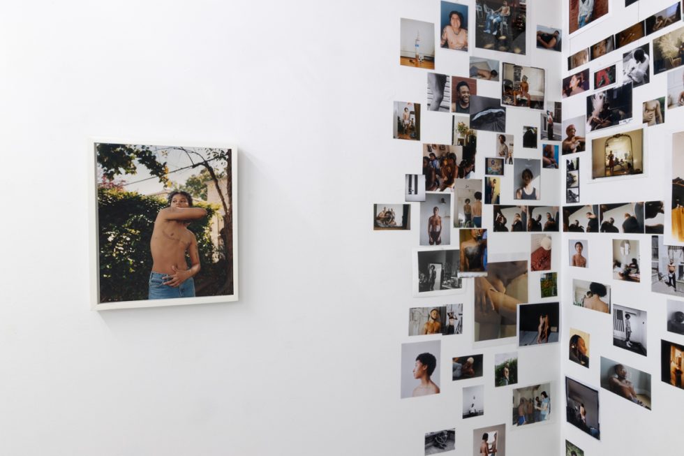 This image shows a collage of images on the walls of a gallery, placed next to a single framed image of a shirtless person with their arm wrapped around their body.