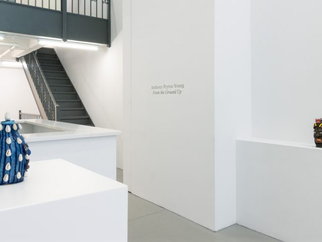 This image depicts the title of an exhibition, posted by the stairs leading up. Two sculptures appear, one on either side of the frame.