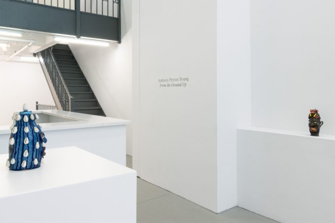 This image depicts the title of an exhibition, posted by the stairs leading up. Two sculptures appear, one on either side of the frame.
