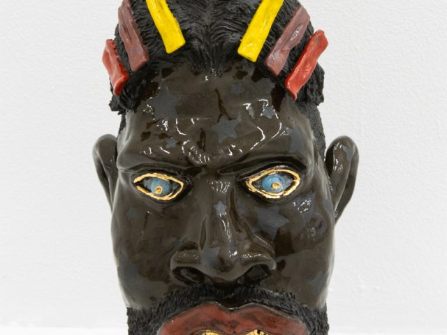 In this image, a ceramic sculpture faces forwards. It's skin is dark brown, and a crown of flowers sits on its hair.