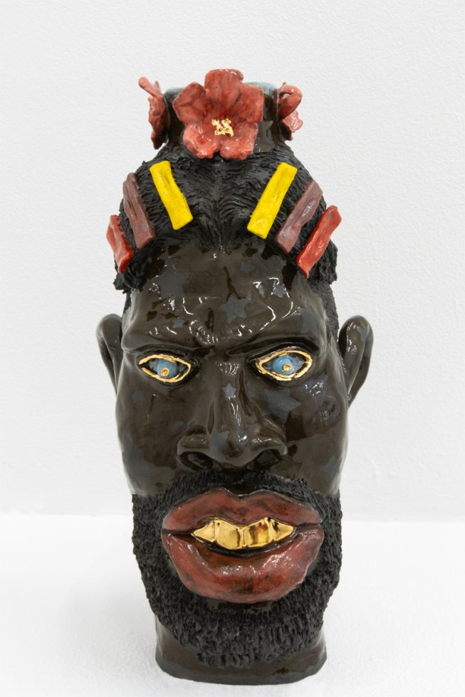 In this image, a ceramic sculpture faces forwards. It's skin is dark brown, and a crown of flowers sits on its hair.