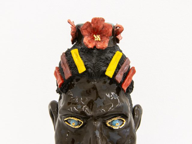 This photograph shows a ceramic sculpture in the shape of a man's head with multi-colored flowers and hair pieces.