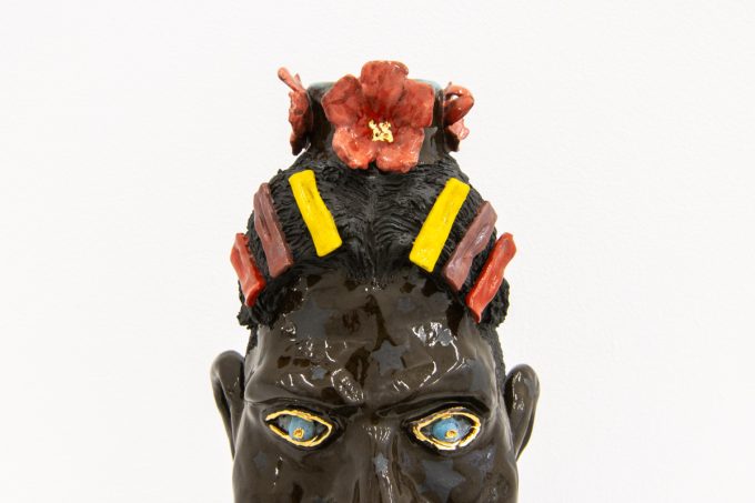 This photograph shows a ceramic sculpture in the shape of a man's head with multi-colored flowers and hair pieces.