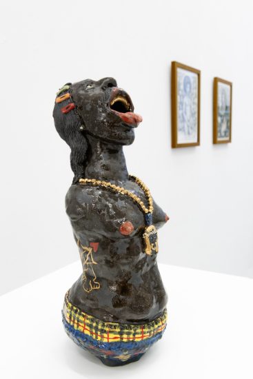 In this image, a ceramic sculpture of a woman with their tongue out leans backwards. A blurry set of framed photographs sits on the walls in the background.