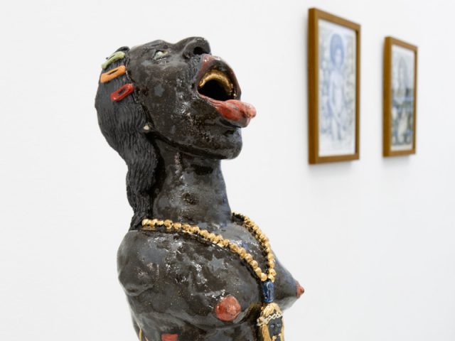 In this image, a ceramic sculpture of a woman with their tongue out leans backwards. A blurry set of framed photographs sits on the walls in the background.