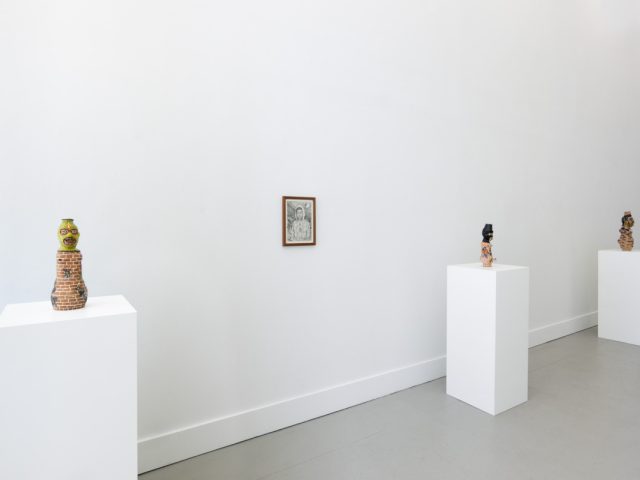 In this photograph, three ceramic sculptures sit on pedestals. Between the first two, a black and white drawing is hung on the wall.