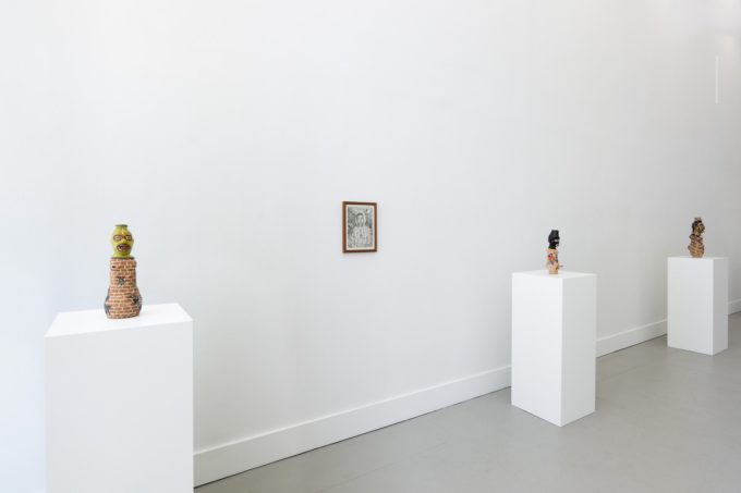 In this photograph, three ceramic sculptures sit on pedestals. Between the first two, a black and white drawing is hung on the wall.