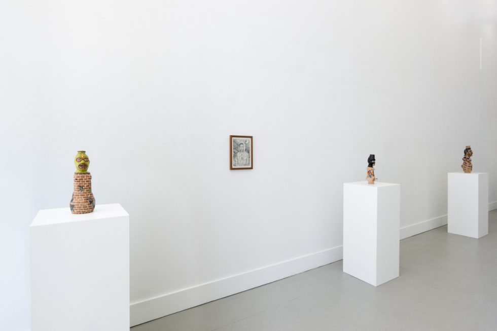In this photograph, three ceramic sculptures sit on pedestals. Between the first two, a black and white drawing is hung on the wall.