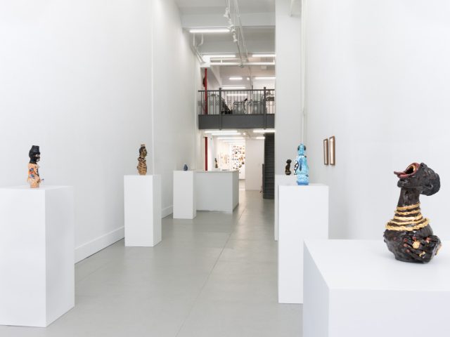 This image shows many ceramic sculptures on pedestals. It looks back towards the end of a gallery, where another room is slightly visible.