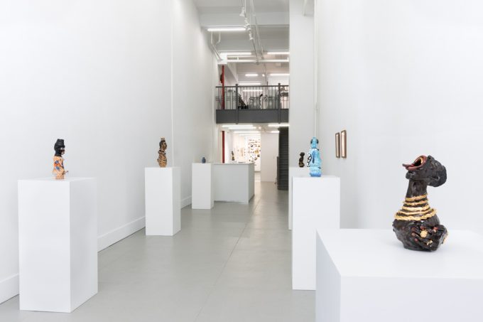 This image shows many ceramic sculptures on pedestals. It looks back towards the end of a gallery, where another room is slightly visible.