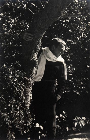In this black and white photograph, a man with a white shirt wrapped around his neck stands in a. forest. One hand holds the trunk of a tree, allowing him to lean out into the trees.