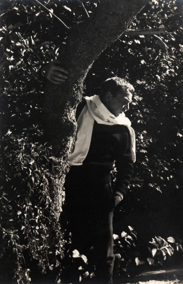 In this black and white photograph, a man with a white shirt wrapped around his neck stands in a. forest. One hand holds the trunk of a tree, allowing him to lean out into the trees.
