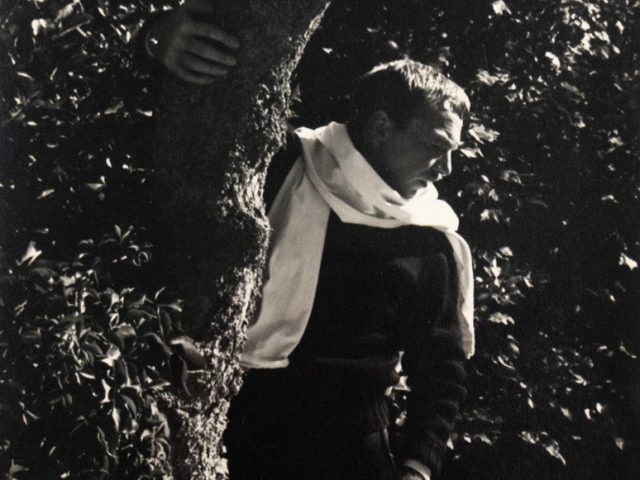 In this black and white photograph, a man with a white shirt wrapped around his neck stands in a. forest. One hand holds the trunk of a tree, allowing him to lean out into the trees.