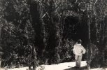 In this black and white photograph, a man wearing white stands on a hill overlooking a forest below him.