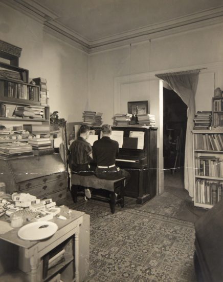In this image, two men sit at a piano. Their backs are facing the camera.