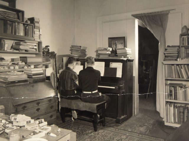 In this image, two men sit at a piano. Their backs are facing the camera.