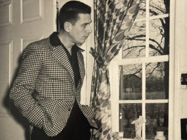 In this image, Paul Cadmus stares out of the window of a white door. His hand is in his pocket.