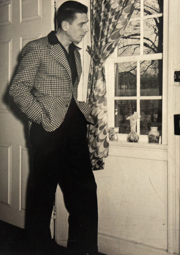 In this image, Paul Cadmus stares out of the window of a white door. His hand is in his pocket.