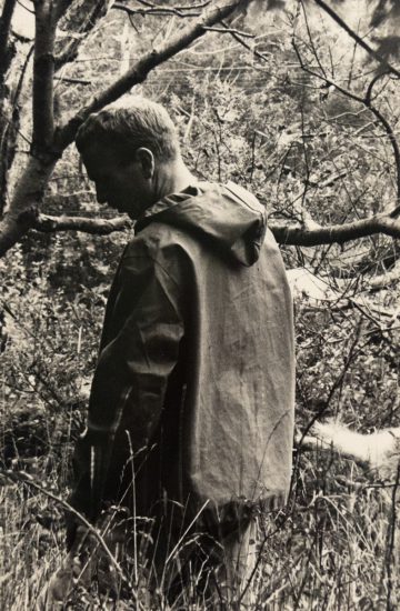In this black and white photograph, a man stands in a forest with his back to the camera. He looks slightly backwards, over his shoulder.