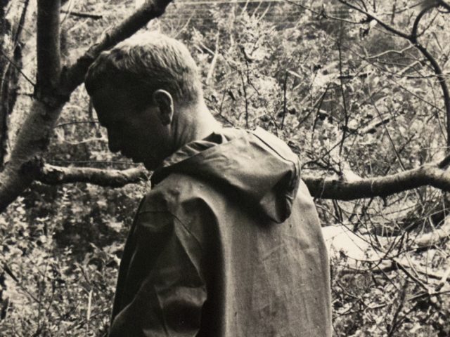 In this black and white photograph, a man stands in a forest with his back to the camera. He looks slightly backwards, over his shoulder.