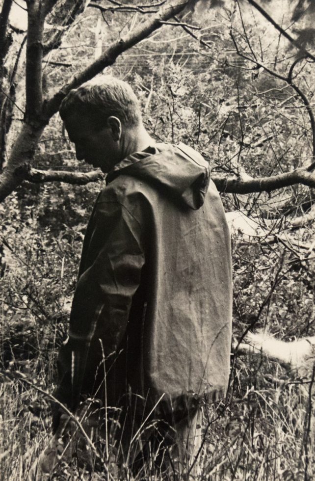 In this black and white photograph, a man stands in a forest with his back to the camera. He looks slightly backwards, over his shoulder.