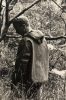 In this black and white photograph, a man stands in a forest with his back to the camera. He looks slightly backwards, over his shoulder.