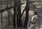 In this black and white photograph, a man leans against a small tree to overlook a river weaving into the distance.